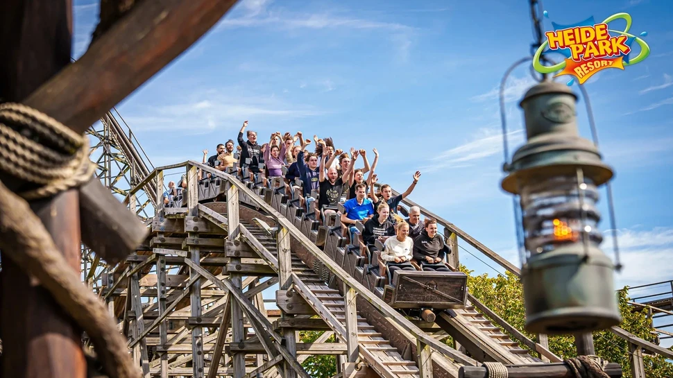 Begeisterte Fahrgäste auf einer Holzachterbahn im Heide Park Resort mit erhobenen Händen bei strahlendem Himmel.
