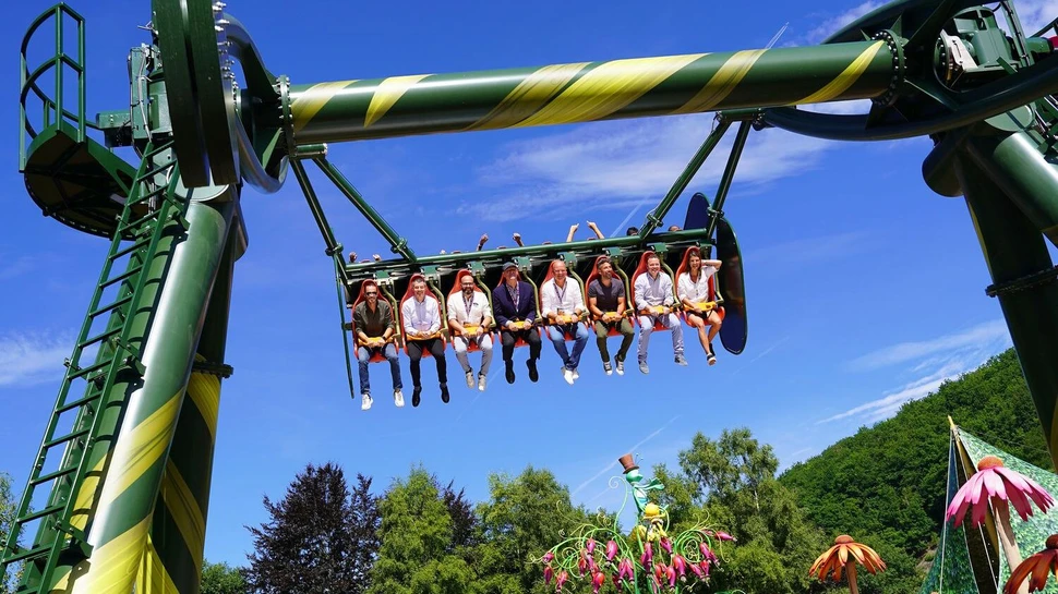 Menschen in einem grün-gelben Fahrgeschäft vor blauem Himmel mit bunten Blumendekoration im Vordergrund.