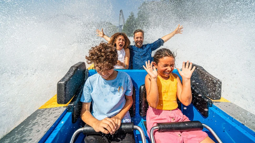 Familie mit zwei Kindern in blauer Wasserbahn, umgeben von spritzenden Wasserfontänen bei sonnigem Wetter.